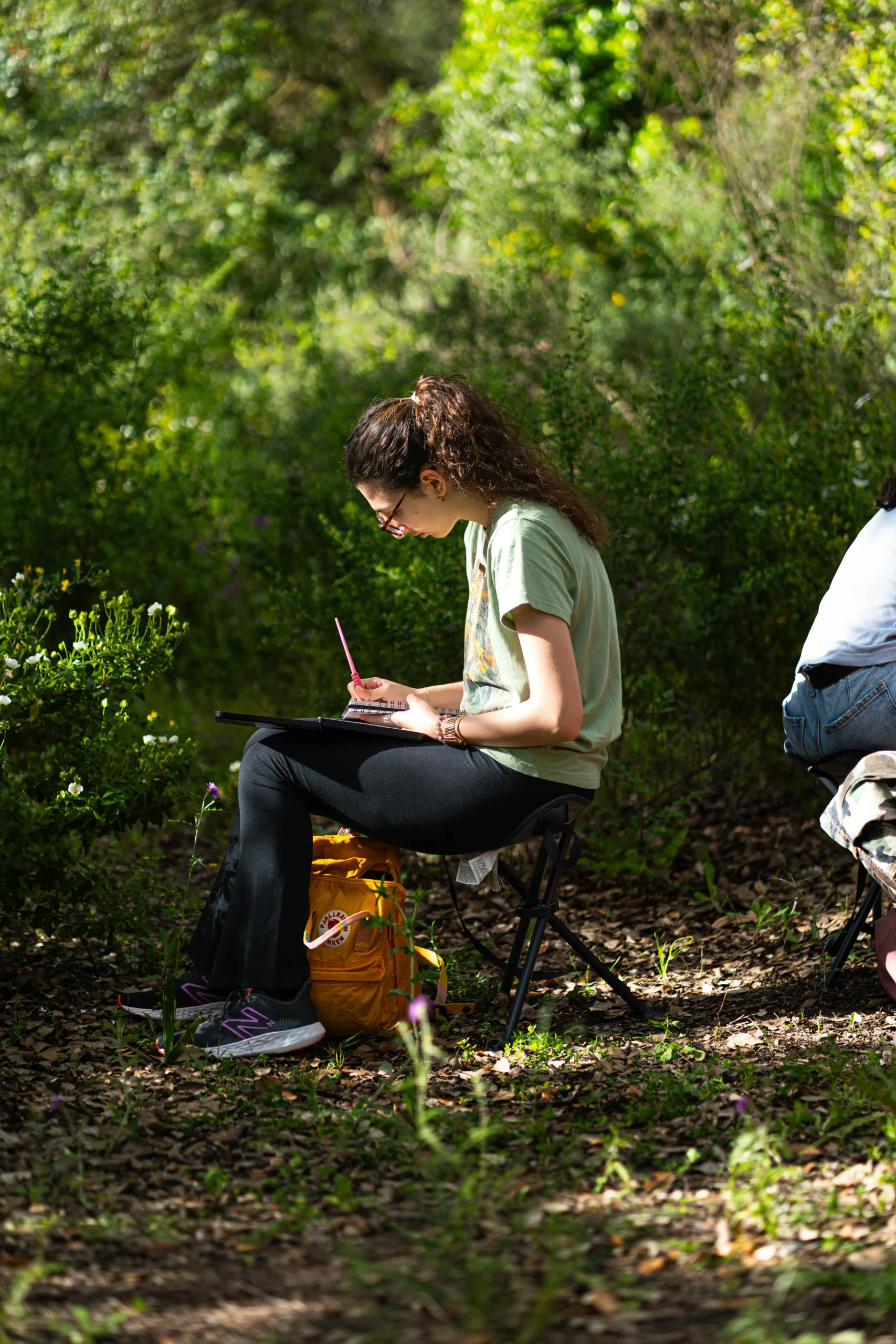Joven con camiseta verde dibujando en un cuaderno sentada en una silla plegable rodeada de vegetación durante una actividad de dibujo al aire libre en las instalaciones de Obimasa, fotografía realizada por DonDigital.