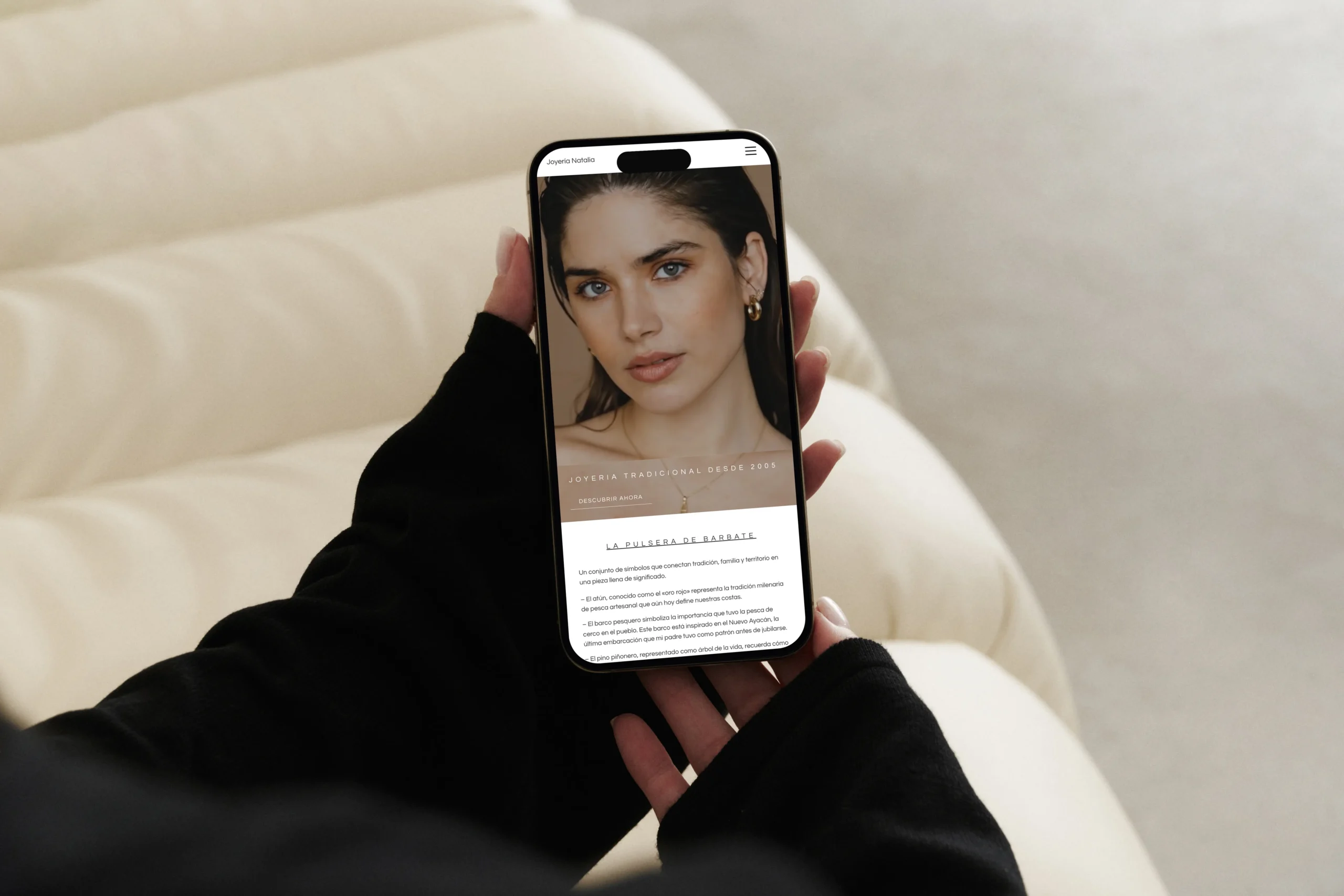 Mano sosteniendo un móvil sobre un sofá beige que muestra la página de inicio y la pulsera de Barbate en la tienda online de Joyería Natalia, fotografía realizada por DonDigital.